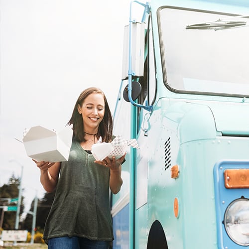 Woman holding to-go food container while walking away from food truck.