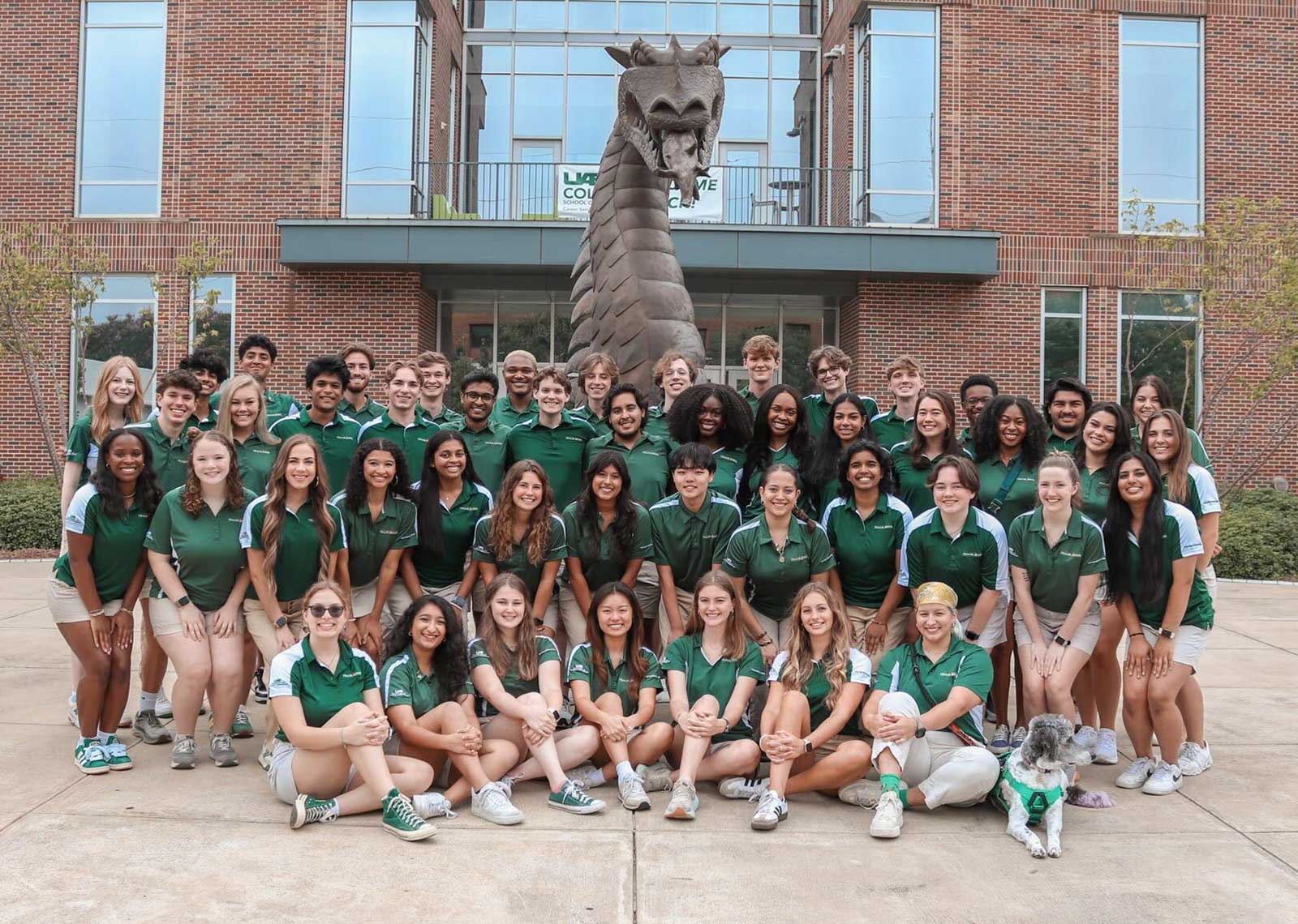 Smiling wearing their official green Trailblazers shirts posing around the bronze Blaze statue outside Collat School of Business.