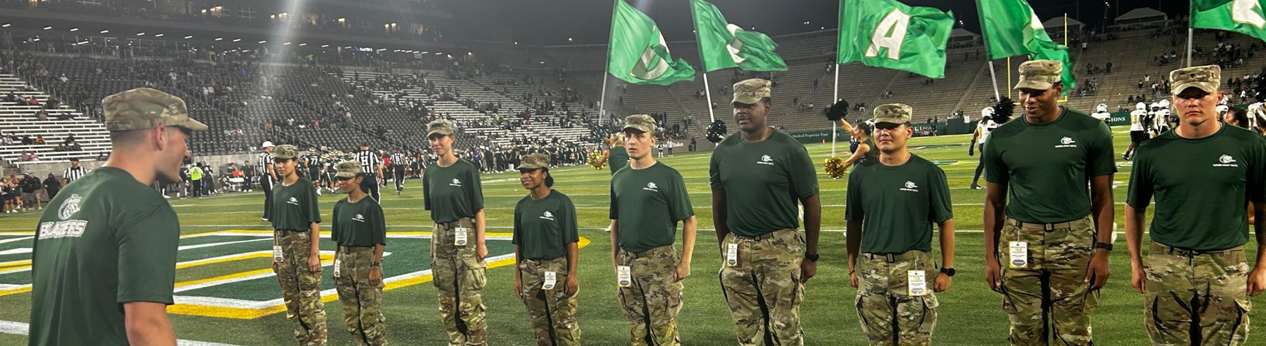 ROTC standing in line on the UAB football field, UAB flags flying behind them.
