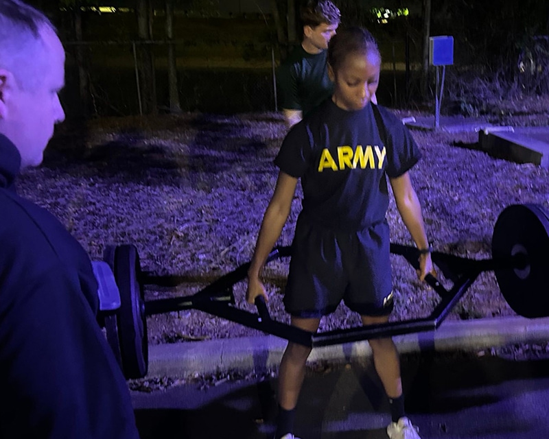 A female student in an army tshirt lifts wight outside at night, supervised by a commander.
