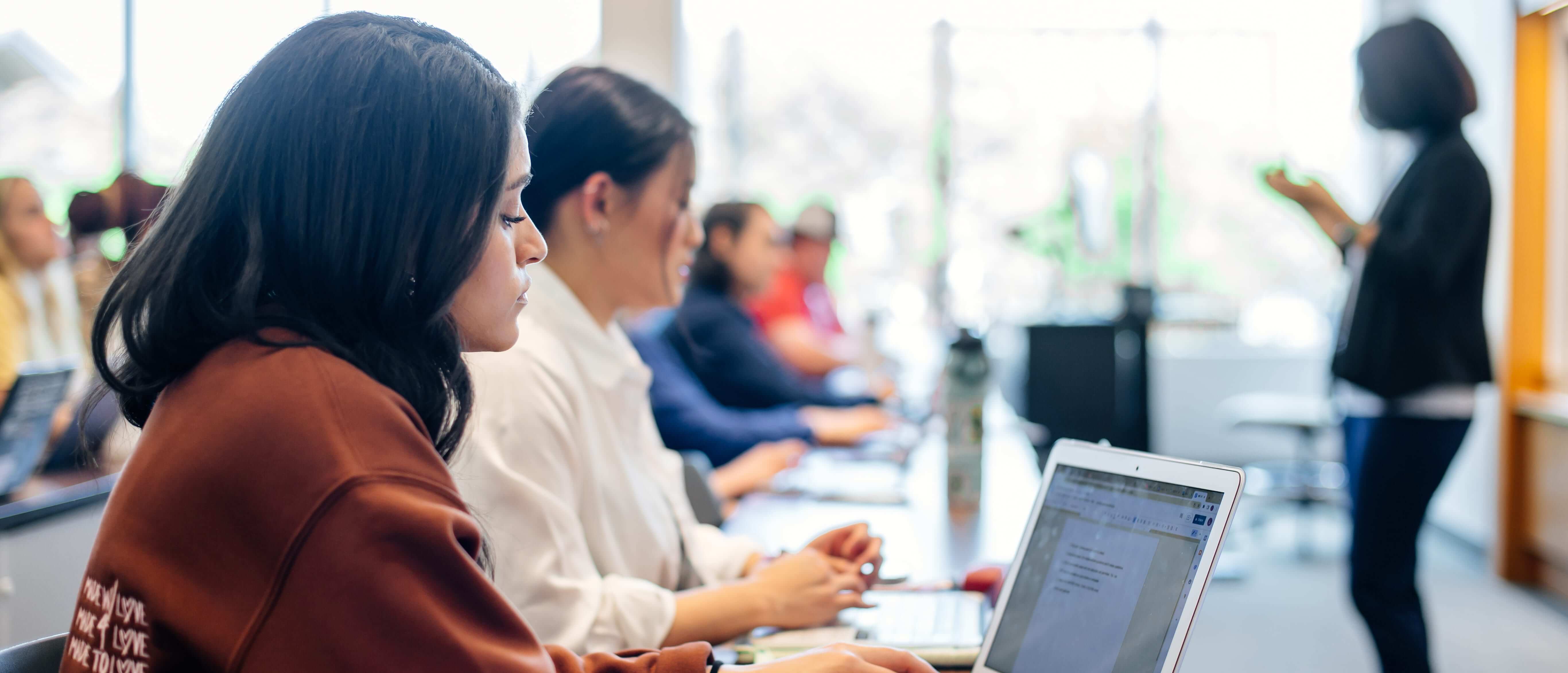 Students in a classroom, a female student looking down at her laptop in the foreground.