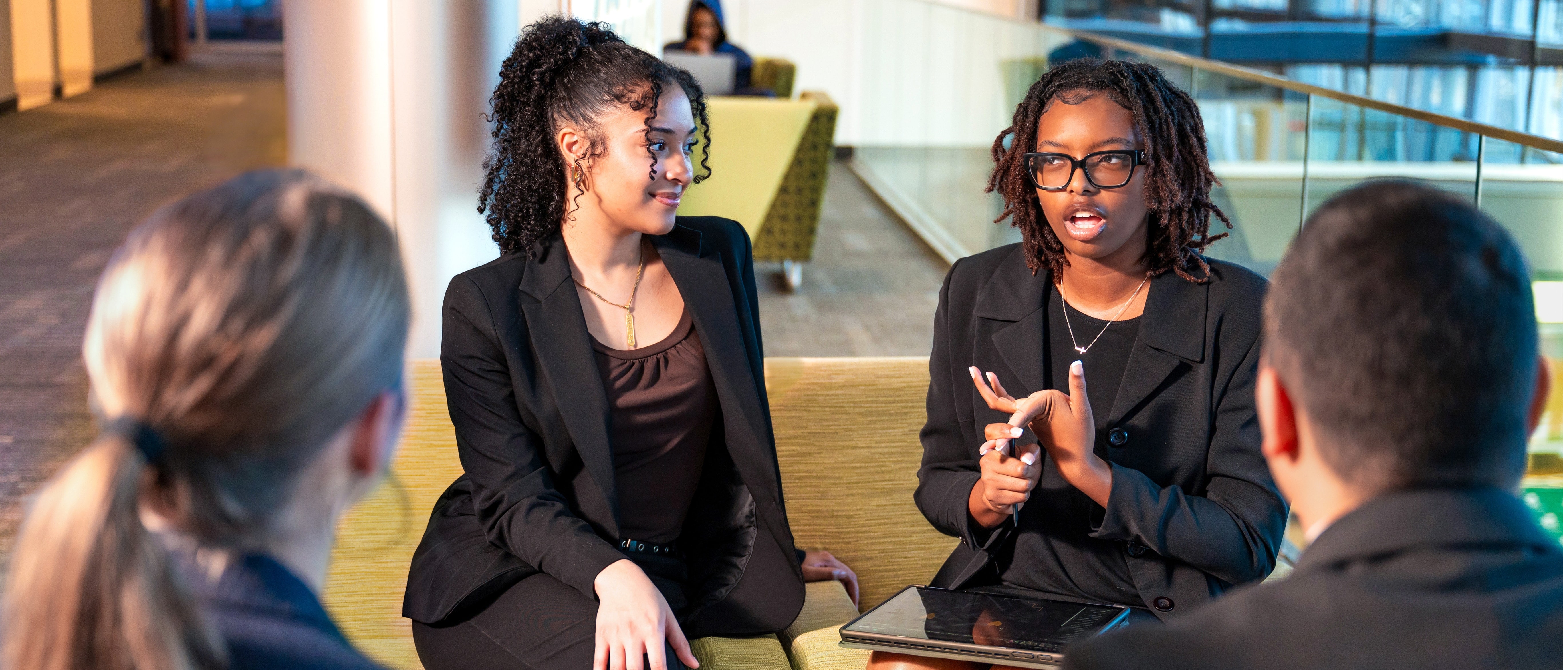 A diverse group of UAB students in black business attire sit on light green couches, listening to one student speak. Two have their backs to the camera.