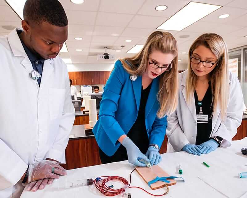 Two students watching a female teacher demonstrate a procedure.