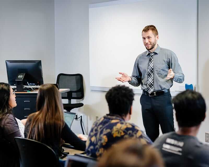 Young male teacher in charcoal shirt and black and white tie in front of a class.