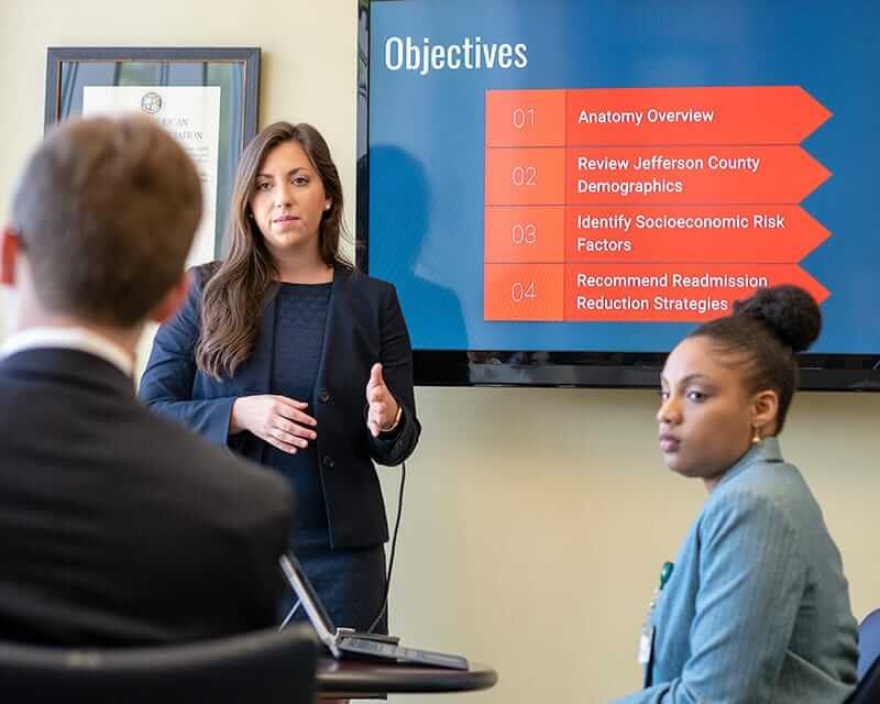 Woman in a blue suit speaking in front of a display with health-related data.