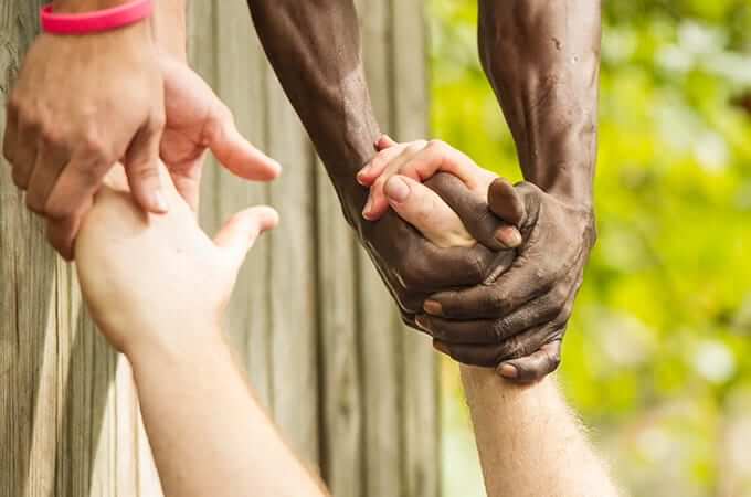 Closeup of hands: two people reaching down to grasp hands of a person attempting to scale a wall.