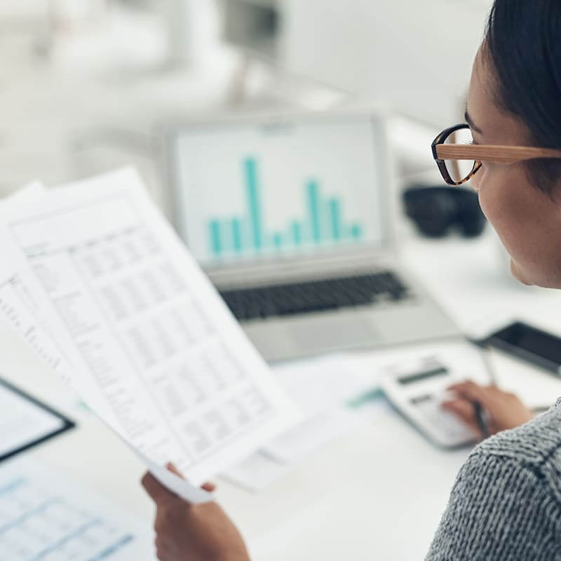 Woman holding a print-out in her left hand and using a calculator with her right hand.