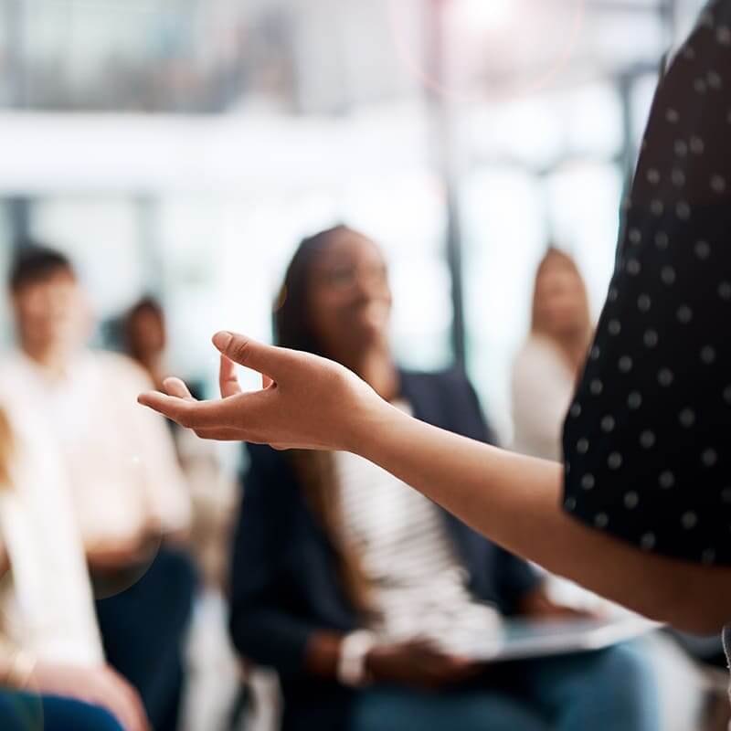 A hand gesturing in the foreground, a group of people out of focus listening to the unpictured presenter.