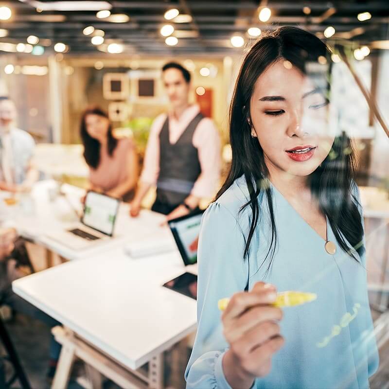 Woman writing on a clear dry-erase board. Behind her a man and a woman at desks are watching her.