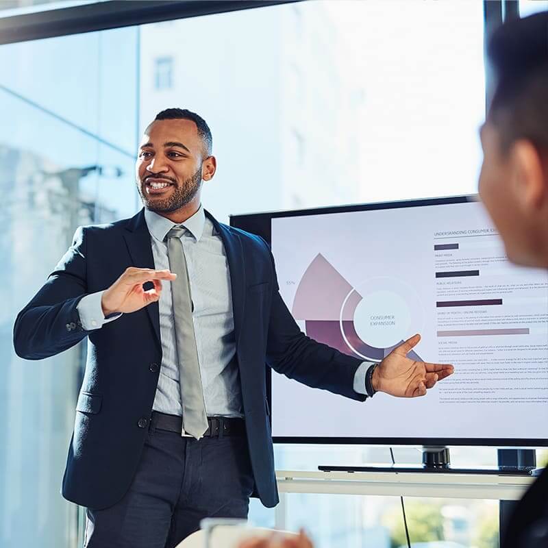 A black man in a suite making a presentation in front of a large screen displaying data.