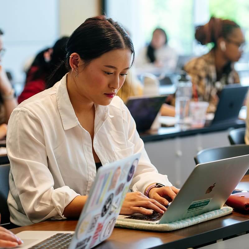 Woman in. a classroom typing on a laptop. 