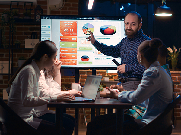 Professional standing in front of coworkers pointing to graphs and charts displayed on widescreen