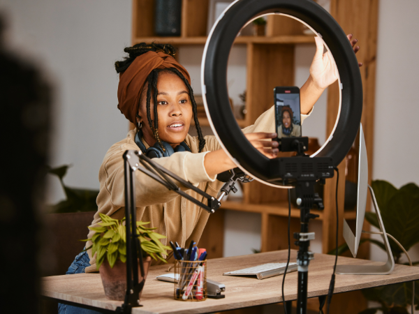 Person recording a video using a smartphone mounted on a ring light, in a room with a plant and artistic supplies.