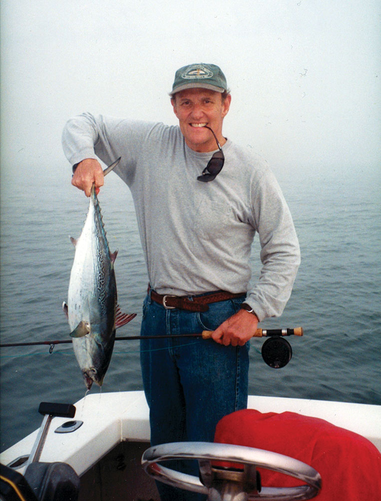 Lee Barton Chapman standing on a boat, holding up a fish, ocean behind him & sunglasses held in his mouth. 