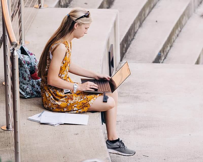 Female student with long blond hair studies outside, wearing a yellow sundress and gray sneakers.