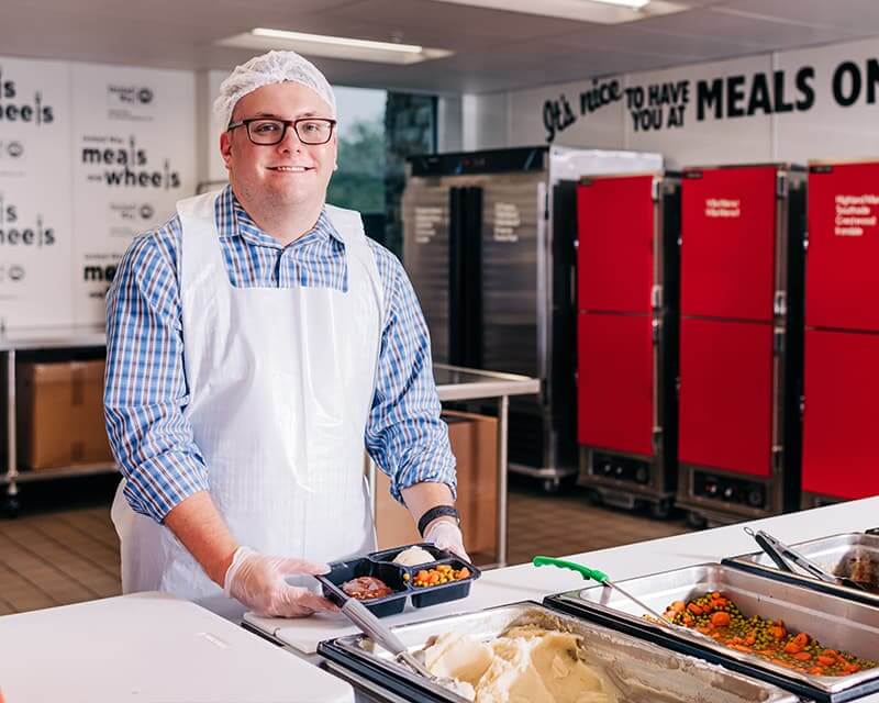 A man wearing glasses, an apron, and hairmask puts together a meal at a Meals on Wheels facility.