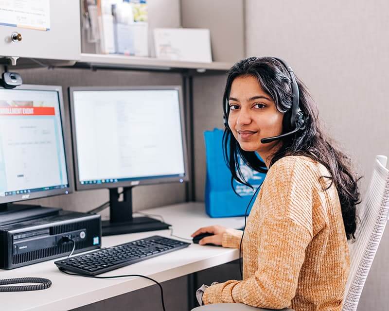 Female student with olive skin and long hair wearing a headset, at a computer in office.