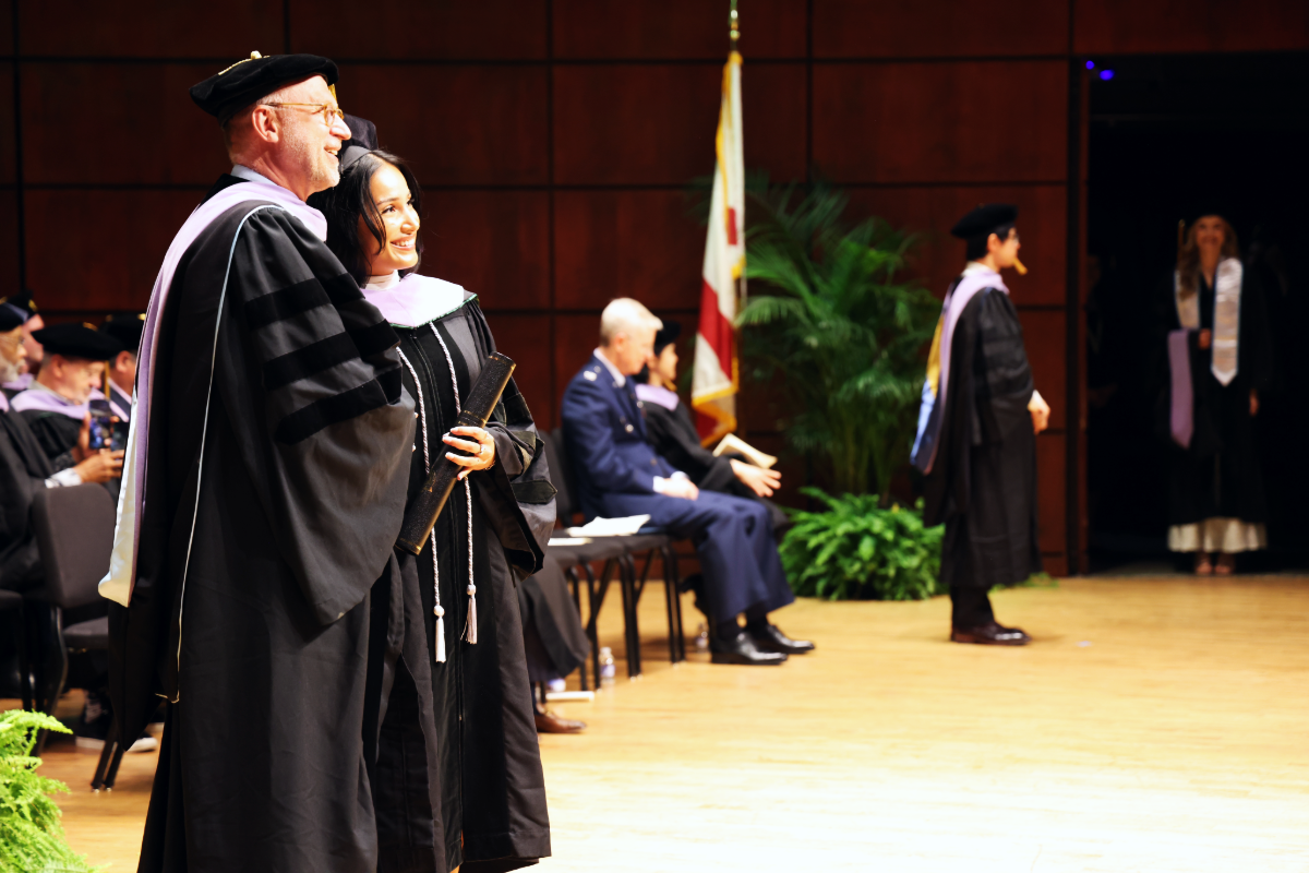 Graduate standing with Dean Geurs on stage at 2026 SOD Commencement Ceremony, faculty members and other graduates in the background
