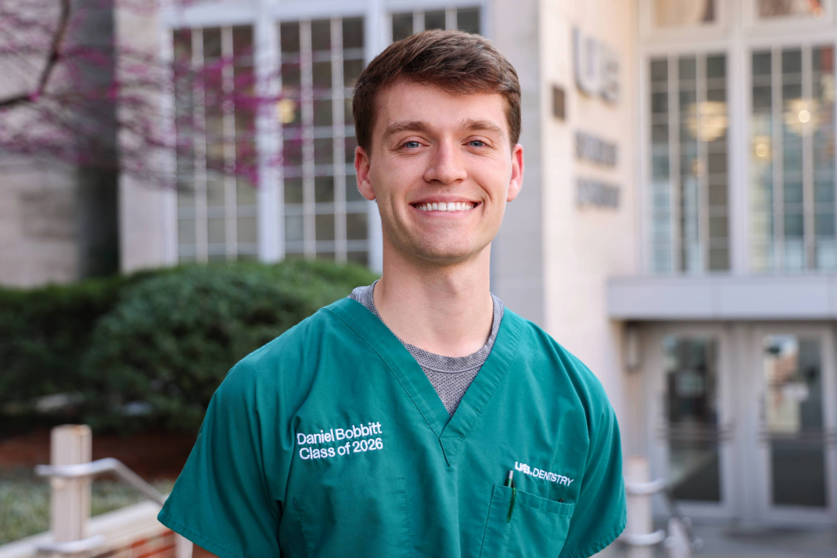 Man in green scrubs labeled, "Daniel Bobbitt Class of 2026 UAB Dentistry" smiling for photo in front of UAB School of Dentistry building.