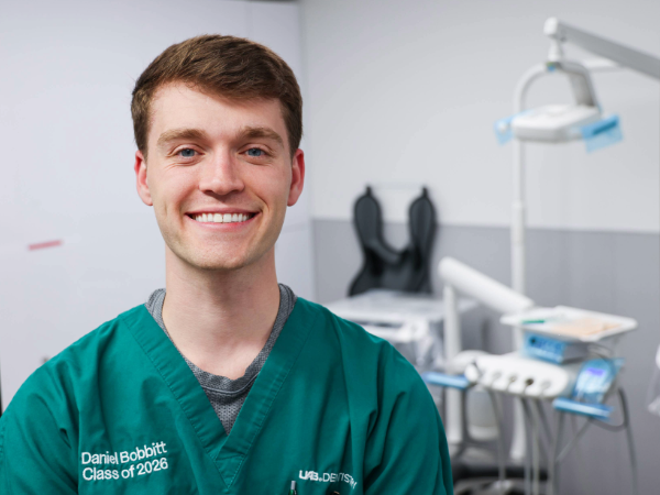 Smiling dental student wearing green scrubs in a clinical training room, with dental equipment and chair visible in the background.