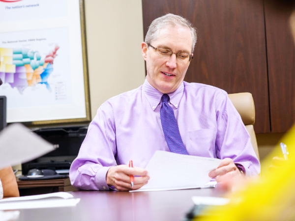 Man in a purple shirt and tie reviewing a document at an office table during a meeting.