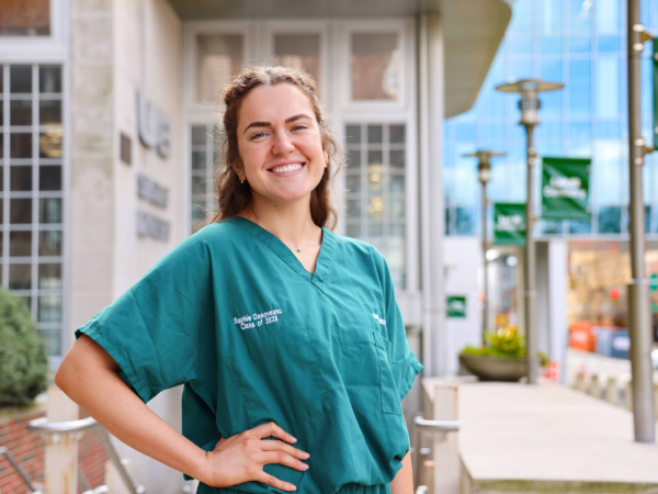 Woman with brown hair and green scrubs smiling with hand on hip while standing outside in front of the UAB School of Dentistry 