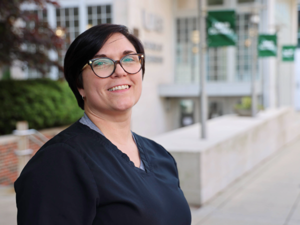 Woman in black medical scrubs with short dark hair and glasses, smiling outdoors.
