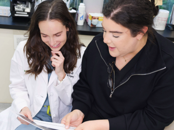 Woman in white coat looking at patient documents next to staff member in black jacket in clinical setting