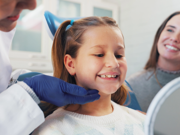 Little girl smiling into mirror at dentist office with dentist and mother in the background