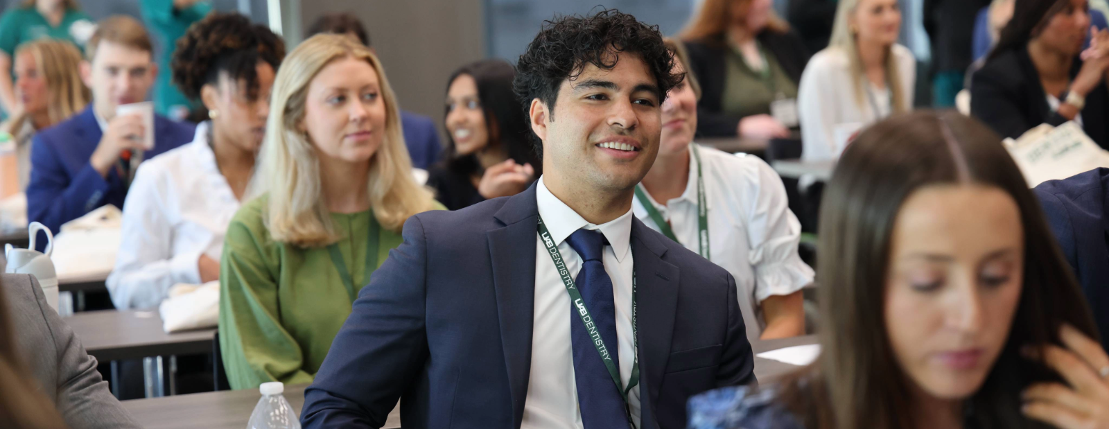 Man with brown hair and blue suit with green lanyard reading "UAB Dentistry" smiling candidly among other sitting in a conference setting