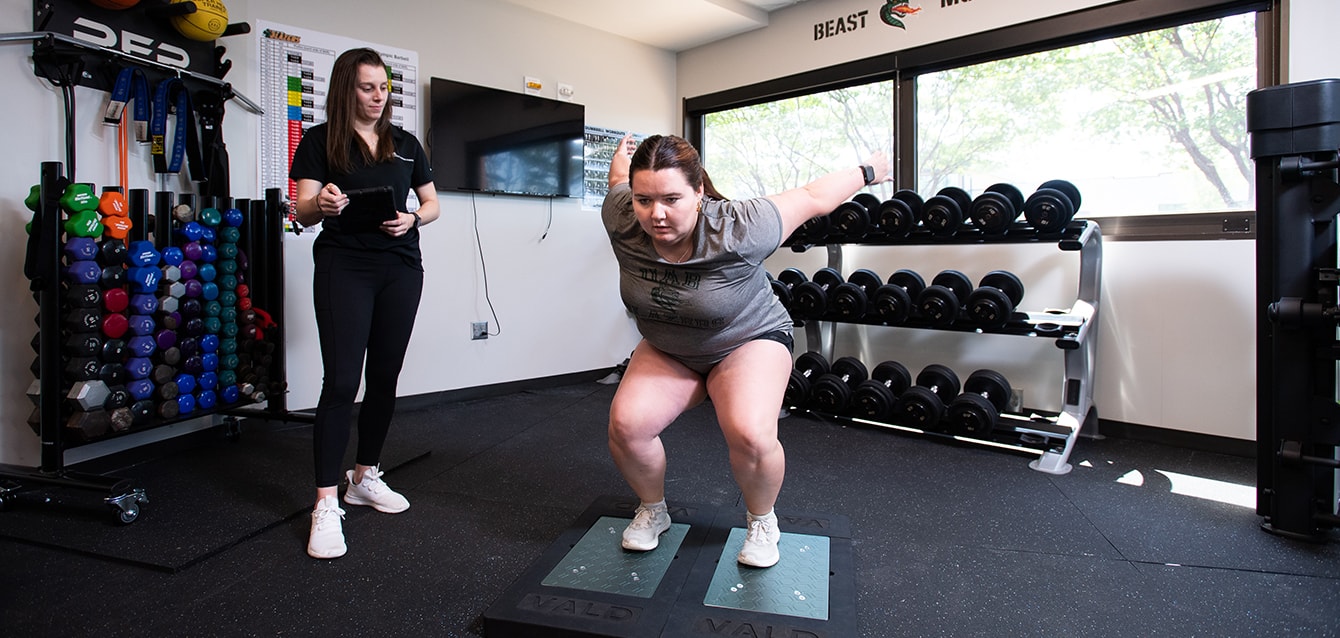 A female trainer in a UAB facility full of hand weights watches a young woman perform a squat.