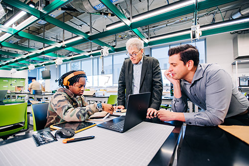 Dr. Timothy Wick, PhD and another man are observing a female engineering student working with a wheelchair ramp prototype in Design x Prototyping Lab in Gorrie Hall.