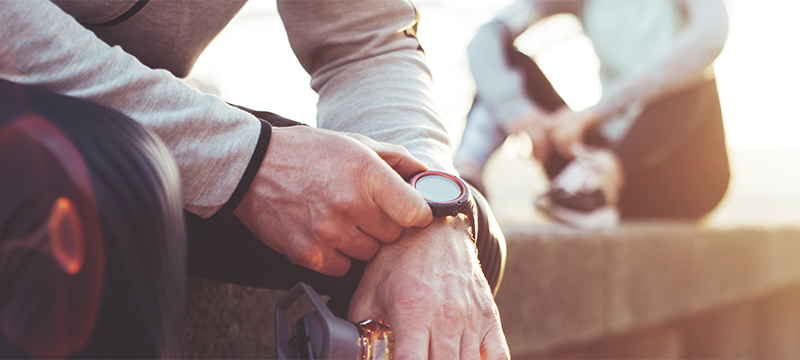 Close-up shot of a man in workout gear adjusting his fitness tracker on his wrist; a woman in the background ties her athletic shoes