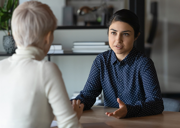 A professionally dressed woman is seen speaking to another woman who is seated across from her at her desk.