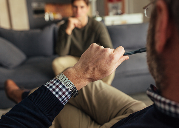 Photo taken over the shoulder of a male therapist chewing on a pen shows his male client out of focus in the background sitting on a couch