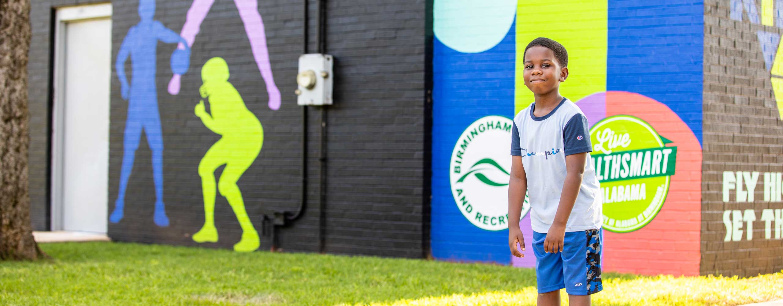 A young Black boy stands outside, in front of a Live Healthsmart mural. 