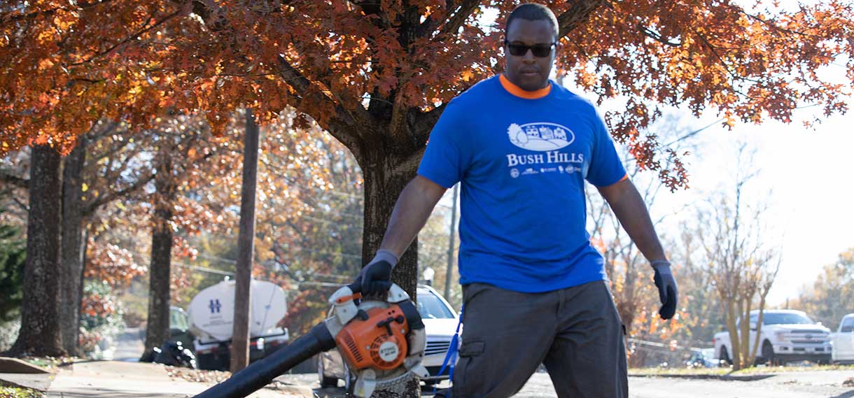 Black male in a blue Bush Hills tshirt carries a chainsaw.