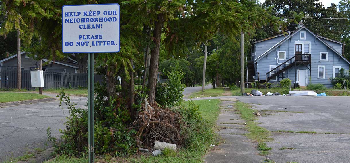 Sign in foreground reads "Help keep our neighborhood clean! Please do not litter!" In the back of the shot, trash is piled near a house.