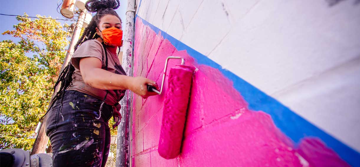 A black woman uses a paint roller to apply bright pink paint to a white wall for the Into The Street Mural.