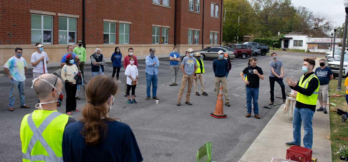 Volunteers stand in a large circle on a street, listening to a man in a safety vest talk.