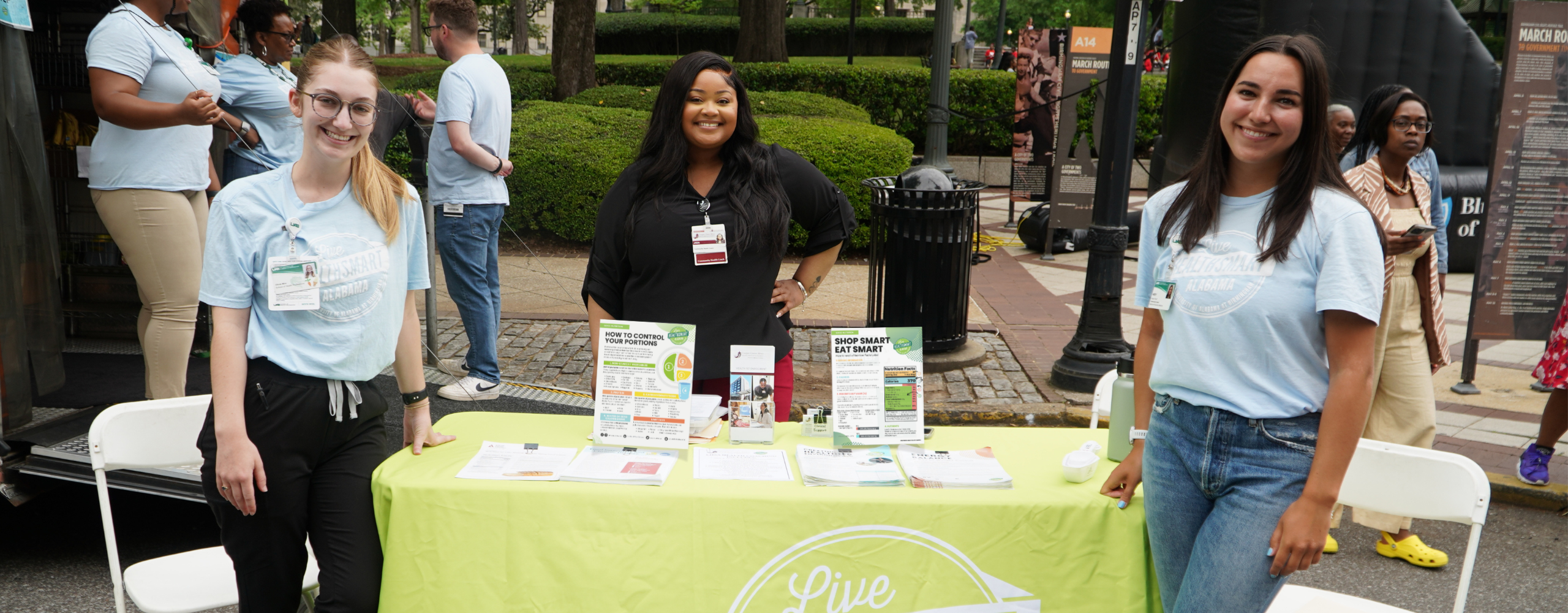 Three female volunteers stand at an info table at an outdoor event.