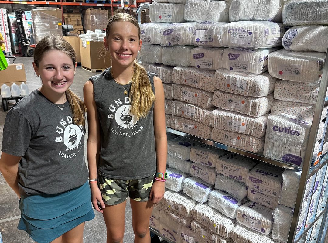 Two young female volunteers stand next to shelves full of bundles.