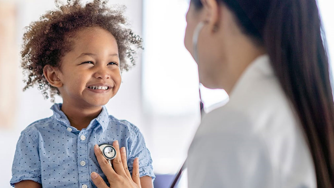 A doctor with long black hair presses a stethoscope against the chest of a smiling young Black boy with exuberant curls. 