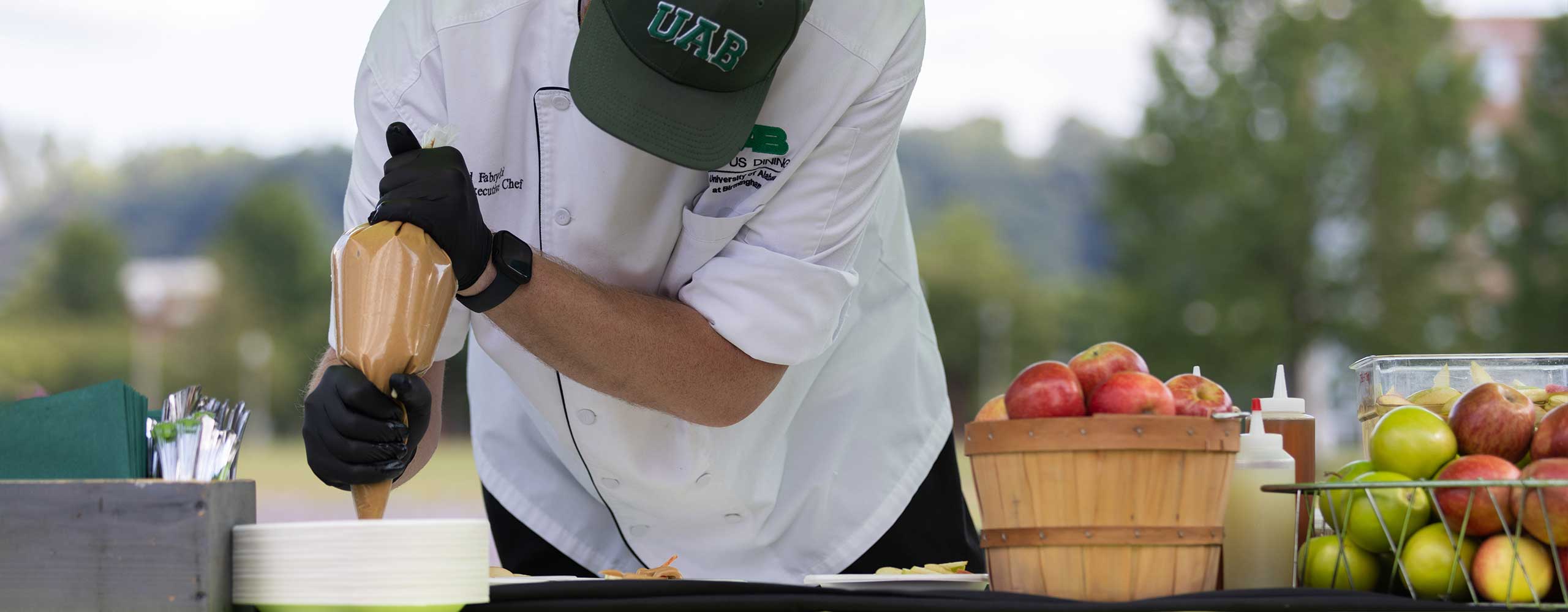 A chef in an outdoor setting and UAB hat uses a pastry bag at a table with a display of fresh fruit. 