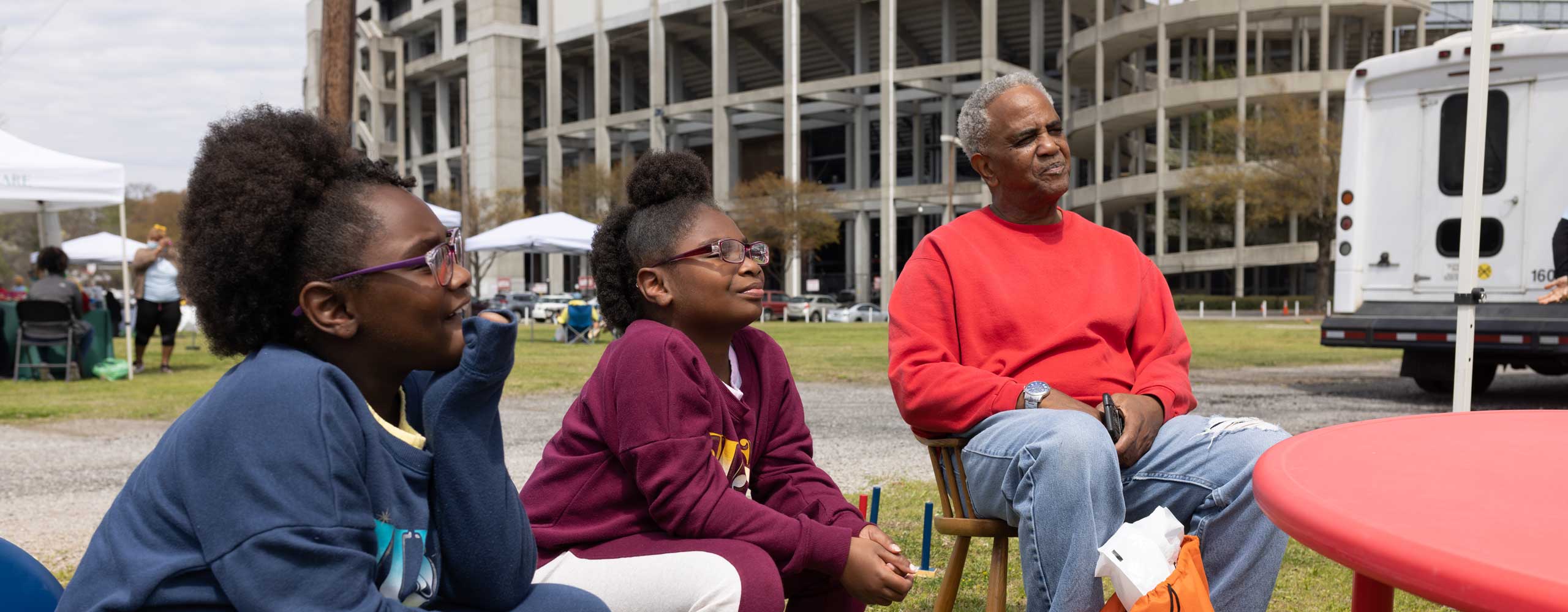 An older black male and two gils sit outside, listening to something, a stadium in the background. 