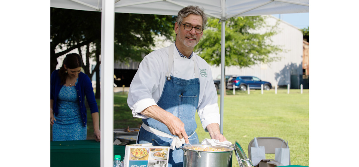 White man with graying hair in glasses and a denim apron points down at a stainless steel pot during cooking demonstration.