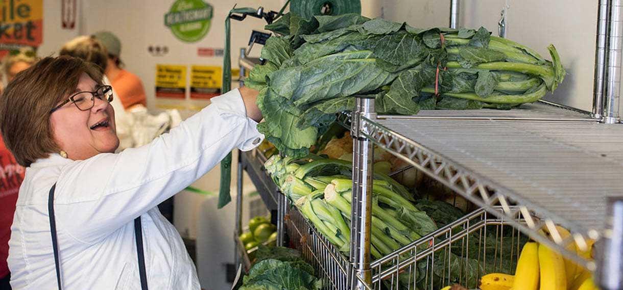 White woman in glasses reaches up happily for some collard greens on a shelf.