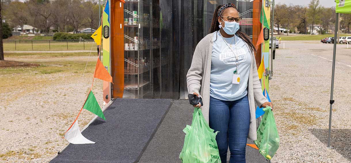 A Black woman leaves the mobile market with several full green grocery bags.