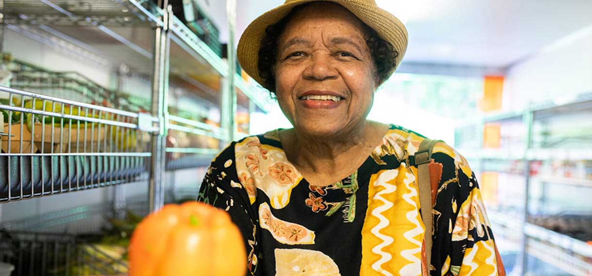 A smiling older black woman in a hat and balck and orange shirt holds up an orange vegetable.