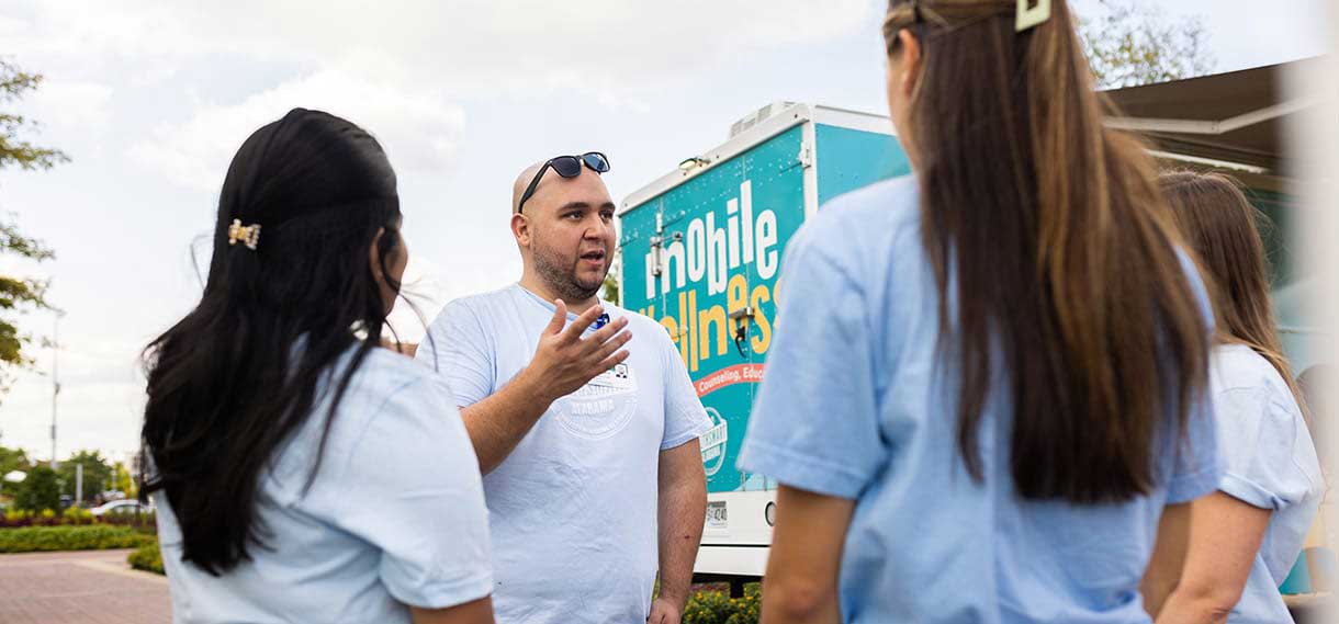A man with sunglasses perched on his head speaks to others outside the mobile wellness van.
